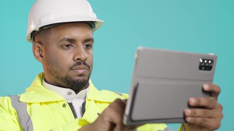 Reviewing plans on a tablet, the concept of engineering and architectural construction, browsing social media, using a modern advanced technical device, a close-up portrait of a Saudi Gulf Arab engineer wearing a jacket and a safety helmet holding a mobile tablet in his hand, green background.