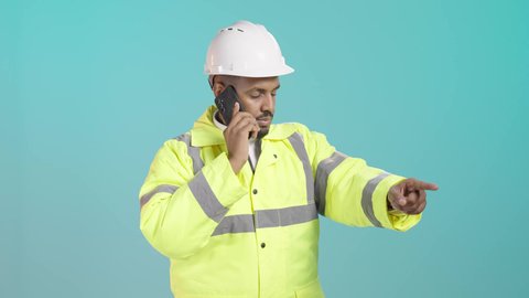 Remote project management, making a phone call via mobile, a portrait of an Arab Gulf Saudi engineer wearing a jacket and safety helmet talking on a mobile phone, pointing to something, green background.