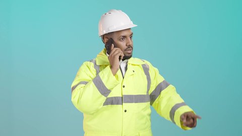 إجراء مكالمه هاتفيه عبرالجوال، إدارة المشاريع عن بعد, بورتريه لمهندس عربي خليجي سعودي يرتدي ستره و خوذة الحماية يتحدث عبر الهاتف المحمول ، الاشارة الى شيء ما ، خلفية خضراء