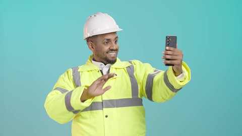 Greeting and welcoming, taking commemorative selfies via mobile, using modern technologies, a portrait of a smiling Saudi Gulf Arab engineer wearing a jacket and safety helmet holding a mobile phone, making a video call via social media applications, green background.