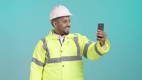 دردشة مرئية عبر الجوال بتعابير الفرح، استخدام الاجهزة التقنية الحديثة، التواصل مع الاهل والاصدقاء، التقاط الصور التذكارية، بورتريه لمهندس عربي خليجي سعودي يرتدي سترة الحماية و الخوذة  يحمل بيده الهاتف المحمول، خلفية خضراء