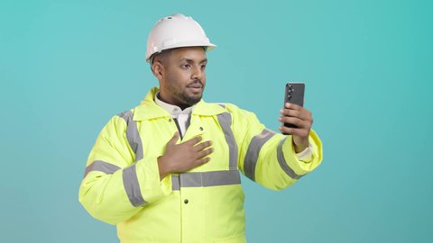 استخدام الاجهزة التقنية الحديثة، التواصل مع الاهل والاصدقاء، التقاط الصور التذكارية، إجراء مكالمة فيديو بتعابير الفرح والسرور، بورتريه لمهندس عربي خليجي سعودي يرتدي سترة الحماية و الخوذة  يحمل بيده الهاتف المحمول، خلفية خضراء