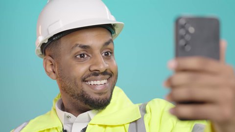 التواصل مع الاهل والاصدقاء، التقاط الصور التذكارية، إجراء مكالمة فيديو بتعابير الفرح والسرور، استخدام الاجهزة التقنية الحديثة، بورتريه مقرب لمهندس عربي خليجي سعودي يرتدي سترة الحماية و الخوذة  يحمل بيده الهاتف المحمول، خلفية خضراء