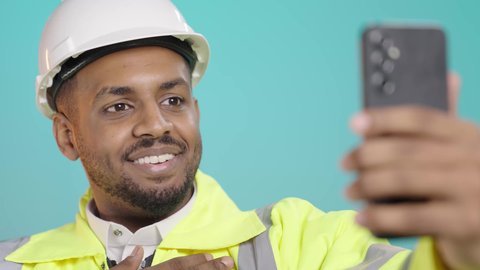 التقاط الصور التذكارية، إجراء مكالمة فيديو بتعابير الفرح والسرور، التواصل مع الاهل والاصدقاء، استخدام الاجهزة التقنية الحديثة، بورتريه مقرب لمهندس عربي خليجي سعودي يرتدي سترة الحماية و الخوذة  يحمل بيده الهاتف المحمول، خلفية خضراء