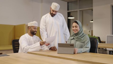 العمل الجماعي و روح الفريق، مفهوم الإدارة المتعاونة، وضع الخطط الاستراتيجية لتحقيق الأهداف، سيدة عربية خليجية عمانية مبتسمة ترتدي عباءة خضراء و حجاب تقوم بمراجعة العمل من خلال التابلت و المناقشة مع زملائها, دراسه المشاريع و مراجعه التقارير السنويه، أداء المهام والاعمال المكتبيه داخل مقر الشركه