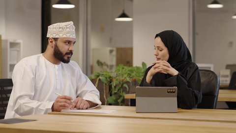 مفهوم الإدارة المتعاونة، تعلم اساليب العمل والتطوير المهني، العمل الجماعي و روح الفريق الواحد، وضع الخطط الاستراتيجية لتحقيق الأهداف،  سيدة عربية خليجية عمانية ترتدي عباءة و حجاب تقوم بمراجعة العمل من خلال التابلت و المناقشة مع زميلها الذي يرتدي الدشداشة و الكمة و يقوم بتدوين الملاحظات