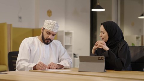 إدارة شؤون الأعمال وتنظيمها، دراسه المشاريع و مراجعه التقارير السنويه، أداء المهام والاعمال المكتبيه داخل مقر الشركه، استخدام التقنيات الحديثة و المتطورة، سيدة عربية خليجية عمانية ترتدي عباءة و حجاب تقوم بمراجعة العمل من خلال التابلت و المناقشة مع زميلها الذي يرتدي الدشداشة و الكمة و يقوم بتدوين الملاحظات