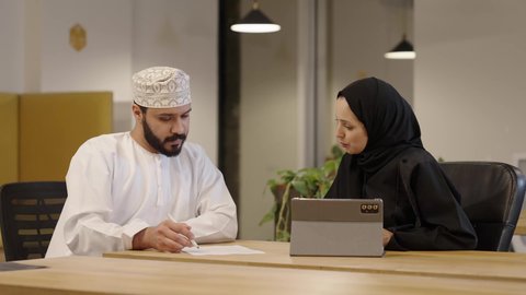 دراسه المشاريع و مراجعه التقارير السنويه، أداء المهام والاعمال المكتبيه داخل مقر الشركه، استخدام التقنيات الحديثة و المتطورة، سيدة عربية خليجية عمانية ترتدي عباءة و حجاب تقوم بمراجعة العمل من خلال التابلت و المناقشة مع زميلها الذي يرتدي الدشداشة و الكمة و يقوم بتدوين الملاحظات