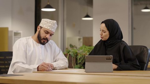 اجتماعات وورشات عمل في مقر الشركة، استخدام أجهزة تقنية حديثة، جمع البيانات وحل المشكلات، سيدة عربية خليجية عمانية ترتدي عباءة و حجاب تقوم بمراجعة العمل من خلال التابلت و المناقشة مع زميلها الذي يرتدي الدشداشة و الكمة و يقوم بتدوين الملاحظات