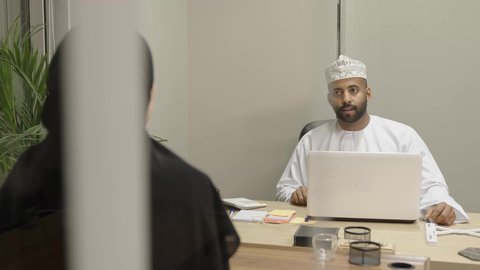 المشاركة في الاراء والمقترحات والحلول, دمج التقنية والتكنولوجيا بالعمل، رجل عربي خليجي عماني يرتدي الدشداشة و الكمة يعمل على اللابتوب و يتناقش مع زميلته التي ترتدي عباءة و حجاب