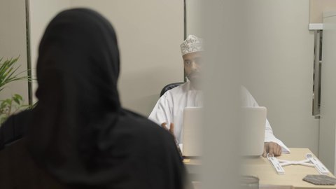 تبادل المعلومات والخبرات, المشاركة في الاراء والمقترحات والحلول, دمج التقنية والتكنولوجيا بالعمل، رجل عربي خليجي عماني يرتدي الدشداشة و الكمة يعمل على اللابتوب و يتناقش مع زميلته التي ترتدي عباءة و حجاب