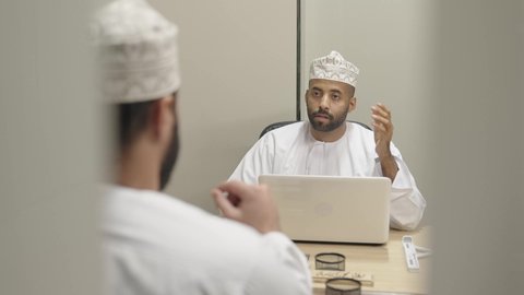 الحوار والنقاش بين الزملاء لإدارة الأعمال وتنظيمها، استخدام التقنيات الحديثة والمطورة، انجاز المشاريع والمهام,  فريق عمل عربي خليجي عماني مكون من رجلين يرتديان الدشداشة والكمة يتناقشان في خطة العمل, إنجاز المهمة المطلوب, تحقيق الاهداف و الاستراتيجيات