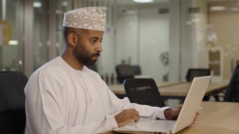 مراجعة الخطط والاستراتيجيات والاتفاقيات عبر التقنيات الحديثه، توفير بيئة عمل مريحة ومناسبة، استخدام جهاز الحاسوب المحمول لإنجاز مهام الاعمال باحترافية، مهن ووظائف ادارية مكتبية, رجل عربي خليجي عماني يرتدي الدشداشة و الكمة يعمل على اللابتوب في المكتب