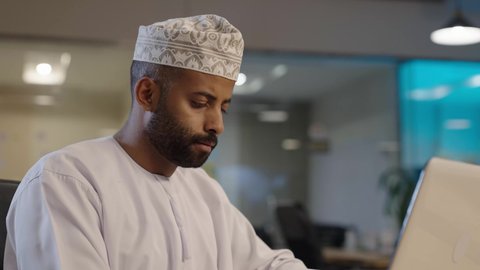 Gestures of focus and seriousness, use of technology and modern techniques, tablet applications and programs, the concept of project management and completing tasks remotely, an Arab Gulf Omani man wearing a dishdasha and a kummah working on a laptop
