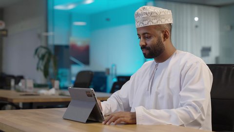 Browsing apps and websites, integrating work with technology, office administrative professions, a smiling Omani Gulf Arab man wearing a dishdasha and a keffiyeh working on a tablet, expressions and gestures of integration