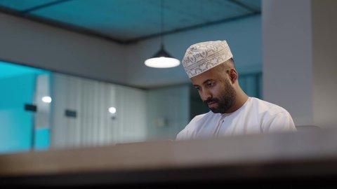 انجاز الأعمال المكتبيه والمهام المطلوبة, رجل عربي خليجي عماني مبتسم يرتدي الدشداشة و الكمة يعمل على التابلت, تعابير وايماءات الاندماج، استخدام التقنية الحديثه المتطورة في الحياة العملية عبر الجهاز اللوحي