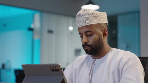 Expressions and gestures of integration, the use of modern advanced technology in practical life via the tablet, office administrative professions, a smiling Arab Gulf Omani man wearing a dishdasha and a turban working on a tablet, browsing applications and websites