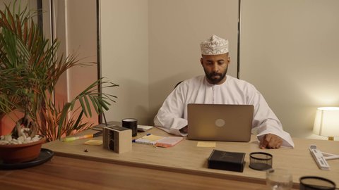 Using computers and technology in organizing and managing businesses, work environment and completing tasks, office administrative professions and jobs, an Arab Gulf Omani man wearing a dishdasha and a kummah working on a laptop in the office