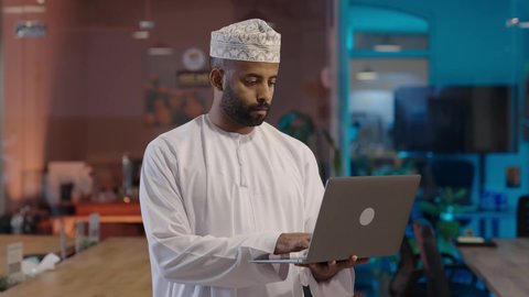 إنجاز المهام العمل المطلوبة, النظر إلى الكاميرا بتعابير السعادة والسرور, إدارة شؤون الأعمال وتنظيمها عبر الاجهزة التقنية، مهن و وظائف ادارية مكتبية, رجل عربي خليجي عماني مبتسم يرتدي الدشداشة و الكمة يعمل على اللابتوب في المكتب