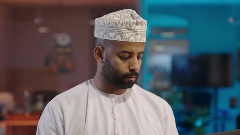 Integrating modern technologies into the administrative and office field, looking at the camera with expressions of happiness and pleasure, managing and organizing business affairs through technical devices, office administrative professions and jobs, an Arab Gulf Omani man wearing a dishdasha and a kummah working on a laptop in the office, completing the required work tasks