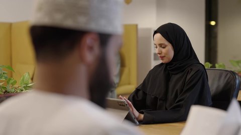 بيئة و مقر العمل, العمل لتحقيق الاهداف و الاستراتيجيات، مهن و وظائف ادارية مكتبية, استخدام الأجهزة التكنولوجية الحديثة والمتطورة، إدارة شؤون الأعمال و تنظيمها، سيدة عربية خليجية عمانية مبتسمة ترتدي حجاب و عباءة تجلس على كرسي مريح و تقوم بالعمل على جهاز التابلت