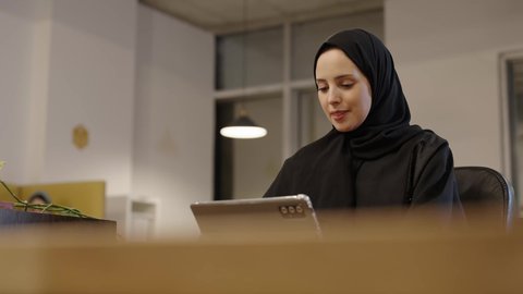 Working to achieve goals and strategies, office administrative professions and jobs, using modern and advanced technological devices, managing and organizing business affairs,  a smiling Omani Gulf Arab woman wearing a hijab and abaya sitting on a comfortable chair and working on a tablet