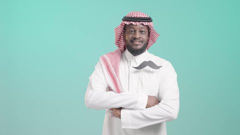 الوقوف باستقامة وأيدي مكتفة، ايماءات الثقة و السرور, الإحتفال باليوم العالمي للرجل 19 نوفمبر، الحفاظ على صحة الذكور، الإهتمام بمظهر الخارجي، بورتريه لرجل أسمر عربي خليجي سعودي يرتدي الثوب التقليدي و الشماغ يضع في جيبه قصاصة ورقية على شكل شارب، خلفية زرقاء