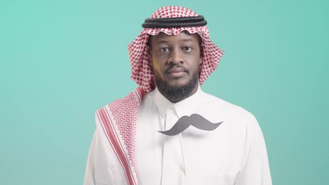 ايماءات الثقة و السرور, الوقوف باستقامة وأيدي مكتفة، الإحتفال باليوم العالمي للرجل 19 نوفمبر، الحفاظ على صحة الذكور، الإهتمام بمظهر الخارجي، بورتريه لرجل أسمر عربي خليجي سعودي يرتدي الثوب التقليدي و الشماغ يضع في جيبه قصاصة ورقية على شكل شارب، خلفية زرقاء