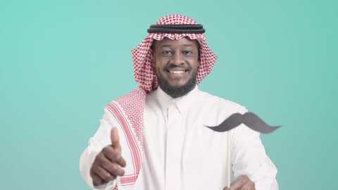 تعابير الرضا و الثقة بالنفس, اليوم العالمي للرجل 19 نوفمبر، بورتريه لرجل أسمر عربي خليجي سعودي يرتدي الثوب التقليدي والشماغ يمسك بيده عود خشبي مع قصاصة ورقية على شكل شارب و يرفع ابهامه بعلامة اللايك، الإهتمام بصحة الرجال، ايماءات السعادة والسرور, خلفية زرقاء