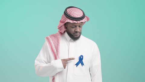 الإشارة الى الشريطة بإيماءات الفرح والفخر، سنوية اليوم العالمي للرجل, بورتريه مقرب لرجل عربي خليجي سعودي يرتدي الثوب التقليدي و الشماغ يشير باصبع السبابة الى شريط أزرق للتوعية بسرطان البروستات، خلفية زرقاء