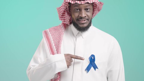 إتباع الإرشادات الطبية وعدم إهمال الصحة الجسدية، أهمية الكشف المبكر عن الأمراض، بورتريه مقرب لرجل عربي خليجي سعودي يرتدي الثوب التقليدي و الشماغ يشير باصبع السبابة الى شريط أزرق للتوعية بسرطان البروستات، خلفية زرقاء