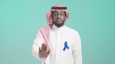 شعار الكشف المبكر عن الأمراض، رفع الكف مفتوحة للتحذير من إهمال الصحة الجسدية، بورتريه مقرب لرجل عربي خليجي سعودي يرتدي الثوب التقليدي و الشماغ  يحمل شريط أزرق للتوعية بسرطان البروستات، الوقوف باستقامة بإيماءات الحزم والجدية، خلفية زرقاء