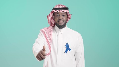 تعابير الإعجاب والموافقة على الكشف المبكر، بورتريه لرجل أسمر عربي خليجي سعودي يرتدي الثوب التقليدي و الشماغ  يضع شريط أزرق للتوعية بسرطان البروستات و يرفع اصبع الابهام باشارة اللايك، خلفية زرقاء