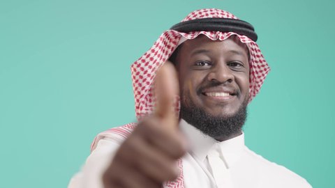 اشارة تعني القبول و السعادة,  بورتريه لرجل أسمر مبتسم عربي خليجي سعودي يرتدي الثوب التقليدي والشماغ يرفع ابهامه بعلامة اللايك، مفهوم النظر إلى الكاميرا بتعابير الرضا، تعبير يدل على الثقة بالنفس, خلفية زرقاء