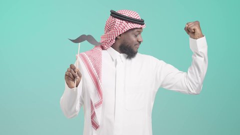 الاهتمام بصحة الذكور، مفهوم القوة والرجولة، النظر إلى الكاميرا بإيماءات الثقة والاعتزاز بالنفس، ذكرى اليوم العالمي للرجل 19 نوفمبر،  بورتريه لرجل أسمر عربي خليجي سعودي يرتدي الثوب التقليدي والشماغ يمسك بيده عود خشبي مع قصاصة ورقية على شكل شارب و يستعرض عضلاته, خلفية زرقاء