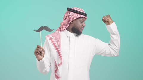 ايماءات قبضة اليد دليل على القوة الجسدية، الإحتفال باليوم العالمي للرجل 19 نوفمبر، مفهوم الرجولة والقوة،  بورتريه لرجل أسمر عربي خليجي سعودي يرتدي الثوب التقليدي والشماغ يمسك بيده عود خشبي مع قصاصة ورقية على شكل شارب و يستعرض عضلاته, خلفية زرقاء