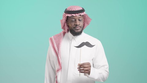 الإهتمام بمظهر الخارجي، اليوم العالمي للرجل 19 نوفمبر، الإحتفال بالمناسبات العالمية، الحفاظ على صحة الذكور، بورتريه لرجل أسمر عربي خليجي سعودي يرتدي الثوب التقليدي و الشماغ يمسك قصاصة ورقية على شكل شارب و يقربها من وجهه، خلفية زرقاء