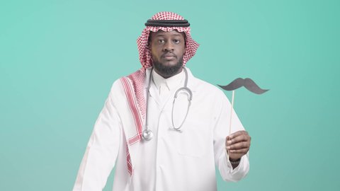 الشعور بالإنزعاج والغضب، رفع اليد بايماءات التحذير والتنبيه، الإهتمام بصحة الذكور، اليوم العالمي للرجل 19 نوفمبر، الإحتفال بالمناسبات العالمية، بورتريه لرجل أسمر عربي خليجي سعودي يرتدي الثوب التقليدي والشماغ و المعطف الطبي والسماعات الطبية يمسك بيده عود خشبي مع قصاصة ورقية على شكل شارب، خلفية زرقاء