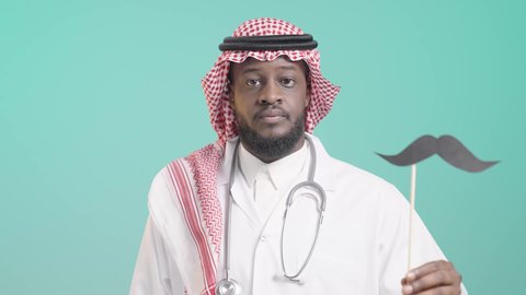 International Men's Day, hand gestures indicating caution and keeping a distance to protect against infection, concept of medicine and healthcare, portrait of a dark-skinned Saudi Arabian Gulf doctor wearing a medical coat and stethoscope holding a wooden stick with a paper clip in the shape of a mustache, blue background