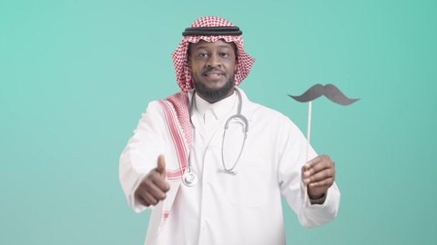 Working in the health sector and the concept of the medical profession, International Men's Day, November 19, portrait of a Saudi Arabian Gulf man wearing a traditional thobe, shemagh, medical coat, and stethoscope, holding a wooden stick with a paper clip in the shape of a mustache and raising his thumb with a like sign, interest in men's health, gestures of happiness and pleasure, blue background