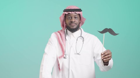 مفهوم مهنة الطب، اليوم العالمي للرجل 19 نوفمبر، بورتريه لرجل عربي خليجي سعودي يرتدي الثوب التقليدي والشماغ و المعطف الطبي و السماعة الطبية يمسك بيده عود خشبي مع قصاصة ورقية على شكل شارب و يرفع ابهامه بعلامة اللايك، الإهتمام بصحة الرجال، ايماءات السعادة والسرور, خلفية زرقاء