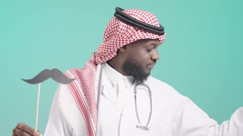 المحافظة على اللياقة والقوة العضلية، اليوم العالمي للرجل 19 نوفمبر، بورتريه لرجل أسمر عربي خليجي سعودي يرتدي الثوب التقليدي والشماغ و المعطف الطبي و السماعة الطبية يمسك بيده عود خشبي مع قصاصة ورقية على شكل شارب و يستعرض عضلاته، الإهتمام بصحة الرجال، ايماءات السعادة والثقة بالنفس, خلفية زرقاء