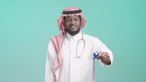 إتباع الأساليب الوقائية, عدم إهمال الصحة الجسدية، أهمية الكشف المبكر عن الأمراض، مفهوم الطب والرعاية الصحية، بورتريه لطبيب أسمر مبتسم عربي خليجي سعودي يرتدي المعطف الطبي والسماعات الطبية يحمل بيده شريط أزرق للتوعية بالأمراض، خلفية زرقاء