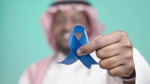 بورتريه مقرب ليد رجل اسمر مبتسم عربي خليجي سعودي يرتدي الثوب التقليدي و الشماغ يمسك بيده شريط ازرق، شعار  اليوم العالمي للرجل، ايماءات السعاده والرضا، خلفيه زرقاء