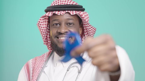 عدم إهمال الصحة الجسدية، أهمية الكشف المبكر عن الأمراض،  مفهوم الطب والرعاية الصحية، بورتريه لطبيب أسمر مبتسم عربي خليجي سعودي يرتدي المعطف الطبي والسماعات الطبية يحمل بيده شريط أزرق للتوعية بالأمراض، خلفية زرقاء