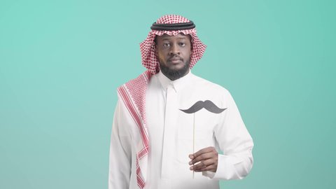 رفع اليد بإيماءات الرفض، اليوم العالمي للرجل 19 نوفمبر، بورتريه لرجل أسمر عربي خليجي سعودي يرتدي الثوب التقليدي و الشماغ يحمل بيده عود خشبي مع قصاصة ورقية على شكل شارب، رفع الكف مفتوحة للتحذير من إهمال الصحة الجسدية، الوقوف باستقامة بإيماءات الحزم والجدية، خلفية زرقاء