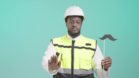 حركات يدين تدل على الأمر بالتوقف عن فعل شيء ما و النهي عنه, الوقوف باستقامة بإيماءات الحزم والجدية، بورتريه لمهندس أسمر عربي خليجي سعودي مبتسم يرتدي الخوذه و سترة الحماية و يمسك بيده عود خشبي مع قصاصة ورقية على شكل شارب و يقوم برفع الكف مفتوحة للتحذير من إهمال الصحة الجسدية، خلفية زرقاء