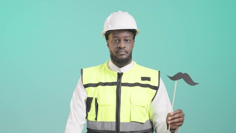 الوقوف باستقامة بإيماءات الحزم والجدية، بورتريه لمهندس أسمر عربي خليجي سعودي مبتسم يرتدي الخوذه و سترة الحماية و يمسك بيده عود خشبي مع قصاصة ورقية على شكل شارب و يقوم برفع الكف مفتوحة للتحذير من إهمال الصحة الجسدية، خلفية زرقاء