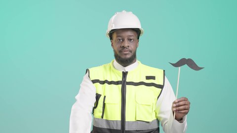 ايماءات الموافقة والإعجاب بشيء ما، ذكرى اليوم العالمي للرجل 19 نوفمبر، بورتريه لمهندس أسمر عربي خليجي سعودي مبتسم يرتدي الخوذه و سترة الحماية و يمسك بيده عود خشبي مع قصاصة ورقية على شكل شارب و يرفع اصبع الابهام بعلامة اللايك, خلفية زرقاء
