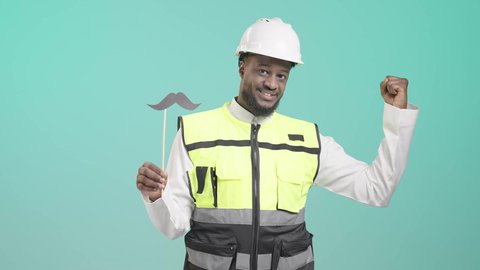 تعابير الرجولة و القوة والثقة بالنفس, اليوم العالمي للرجل 19 نوفمبر، العمل في القطاع الهندسي، بورتريه لمهندس أسمر عربي خليجي سعودي مبتسم يرتدي الخوذه و سترة الحماية و يمسك بيده عود خشبي مع قصاصة ورقية على شكل شارب  و يستعرض عضلاته، خلفية زرقاء